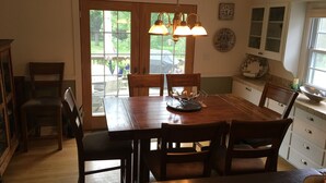 Uma sala de jantar com mesa de madeira, cadeiras e um candelabro. Há janelas com vista para o exterior.