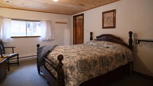 Een slaapkamer met een houten bed, een stoel, een bureau, een raam met gordijnen, een deur en een wandplank.