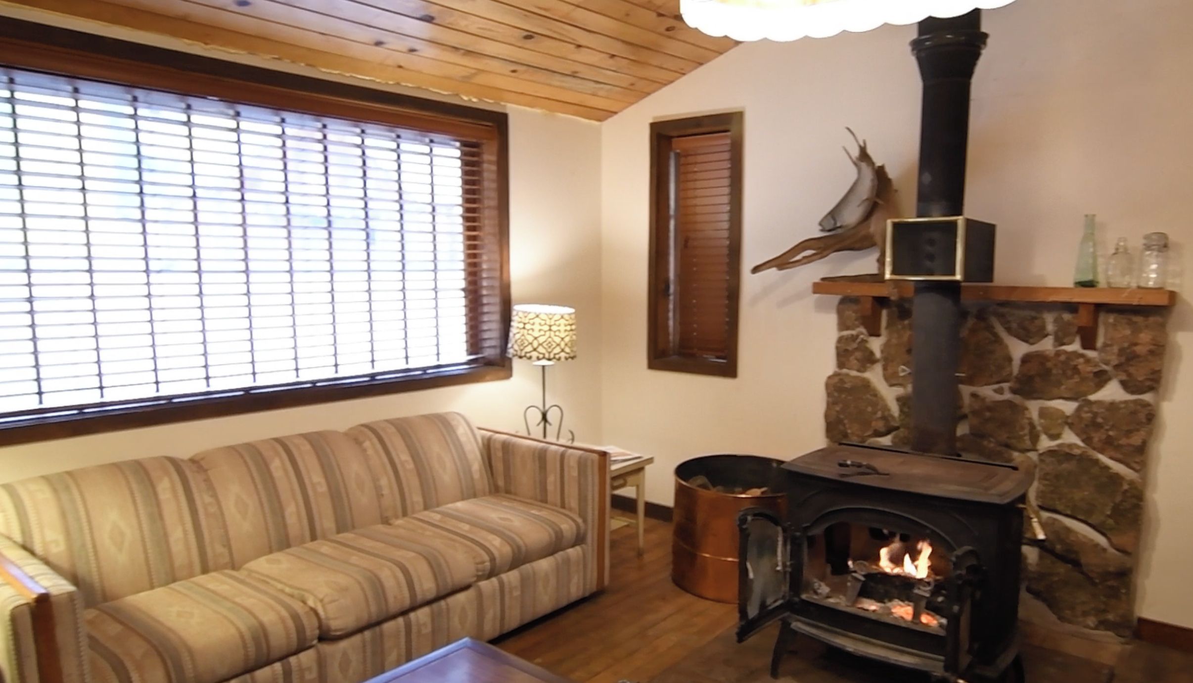 Ein gemütliches Wohnzimmer mit Sofa, Couchtisch, Kamin und Steinwand, sowie ein Fenster mit Jalousien.