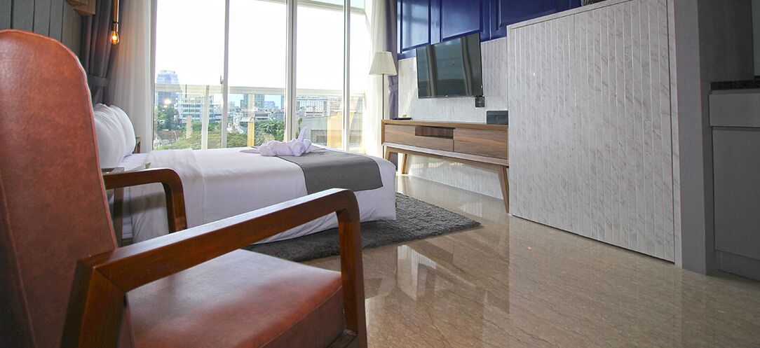 A hotel room with a bed, a desk, a chair, a TV, and a large window offering a city view.