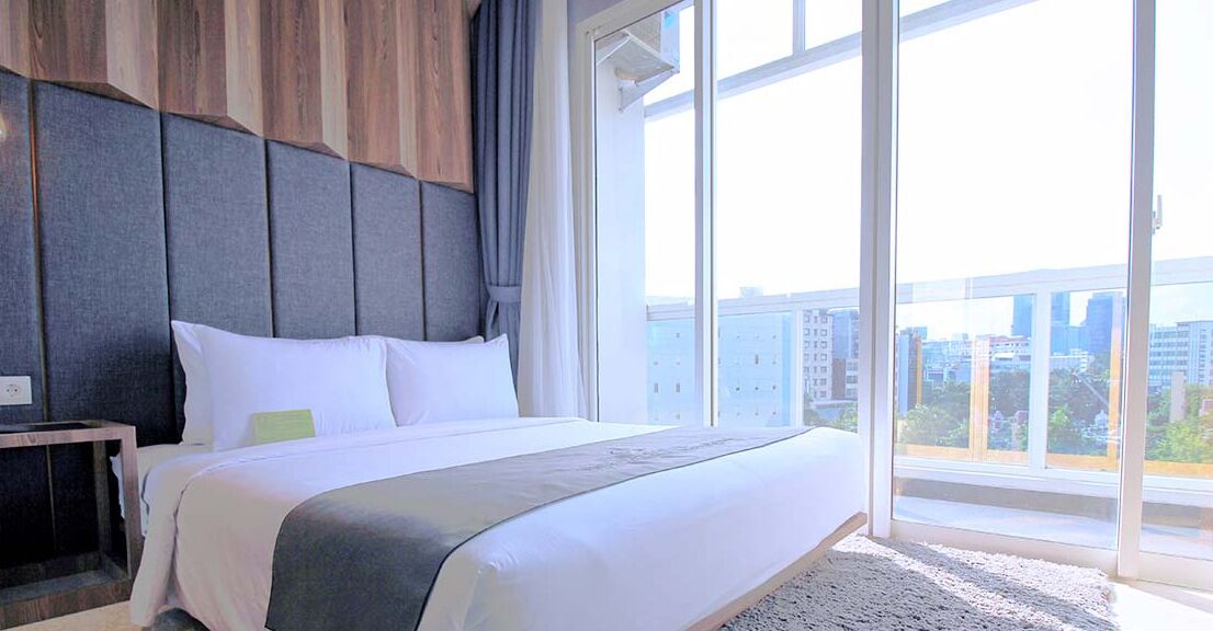 Habitación de hotel con una cama grande, cabecera de madera y vistas de la ciudad a través de amplias ventanas.