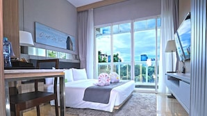 Habitación de hotel con cama, escritorio, televisor y ventana grande con vistas a edificios y zonas verdes.