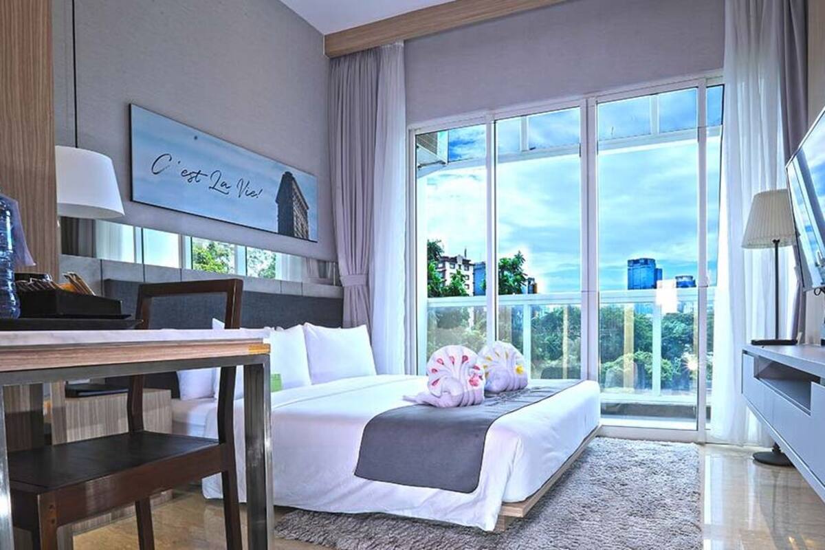 Ein Hotelzimmer mit Bett, Schreibtisch, Fernseher und einem großen Fenster mit Blick auf Gebäude und Grünflächen.