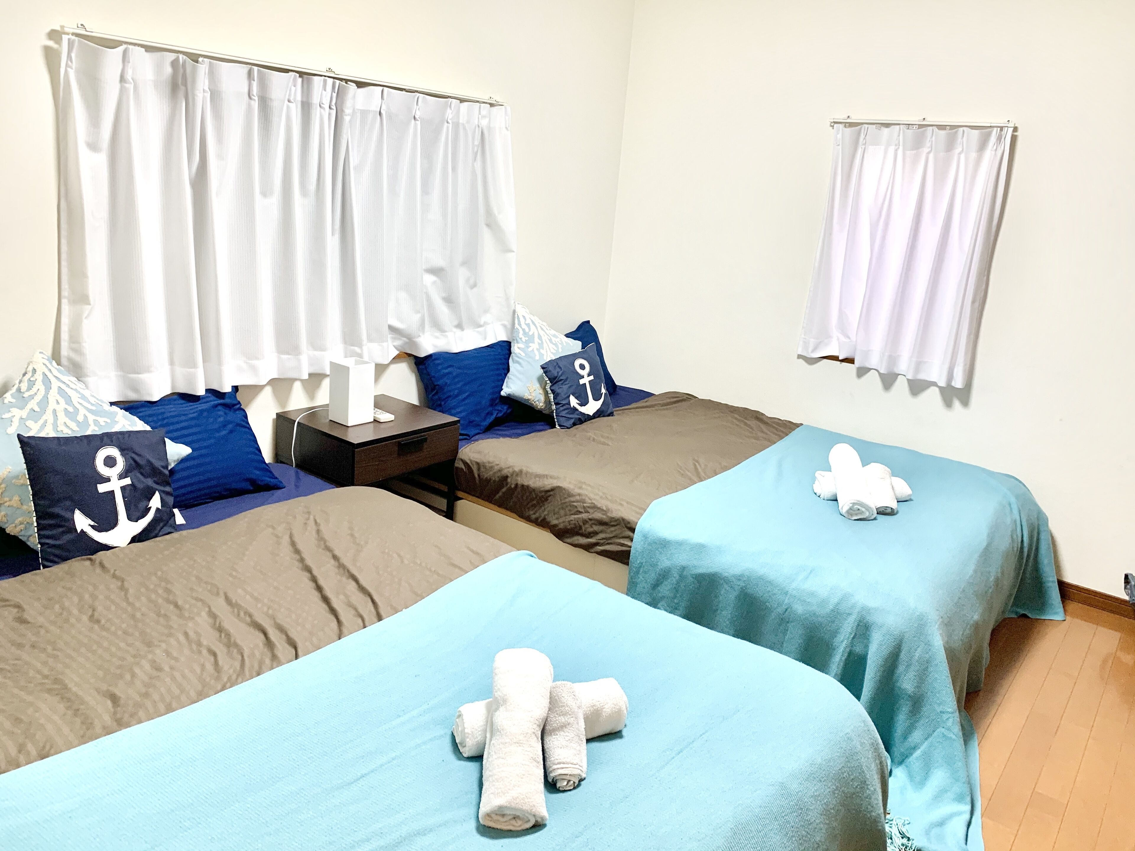Quarto com duas camas, cada uma com colcha azul e toalhas brancas. Cortinas brancas nas janelas, e uma pequena mesa com uma caixa branca e controle remoto.