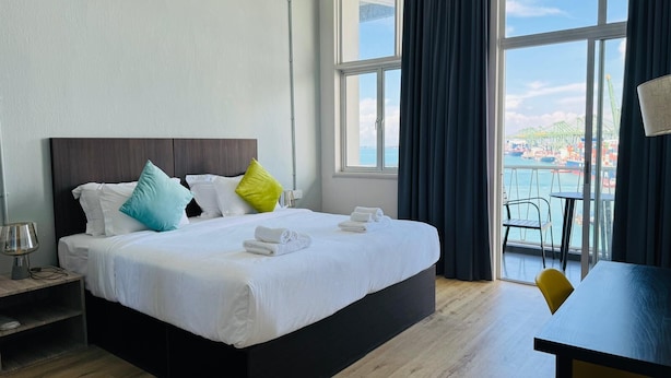 A hotel room with a large bed, a nightstand, a table, and a view of the sea through large windows. - Cooliv Waterfront (Singapore)
