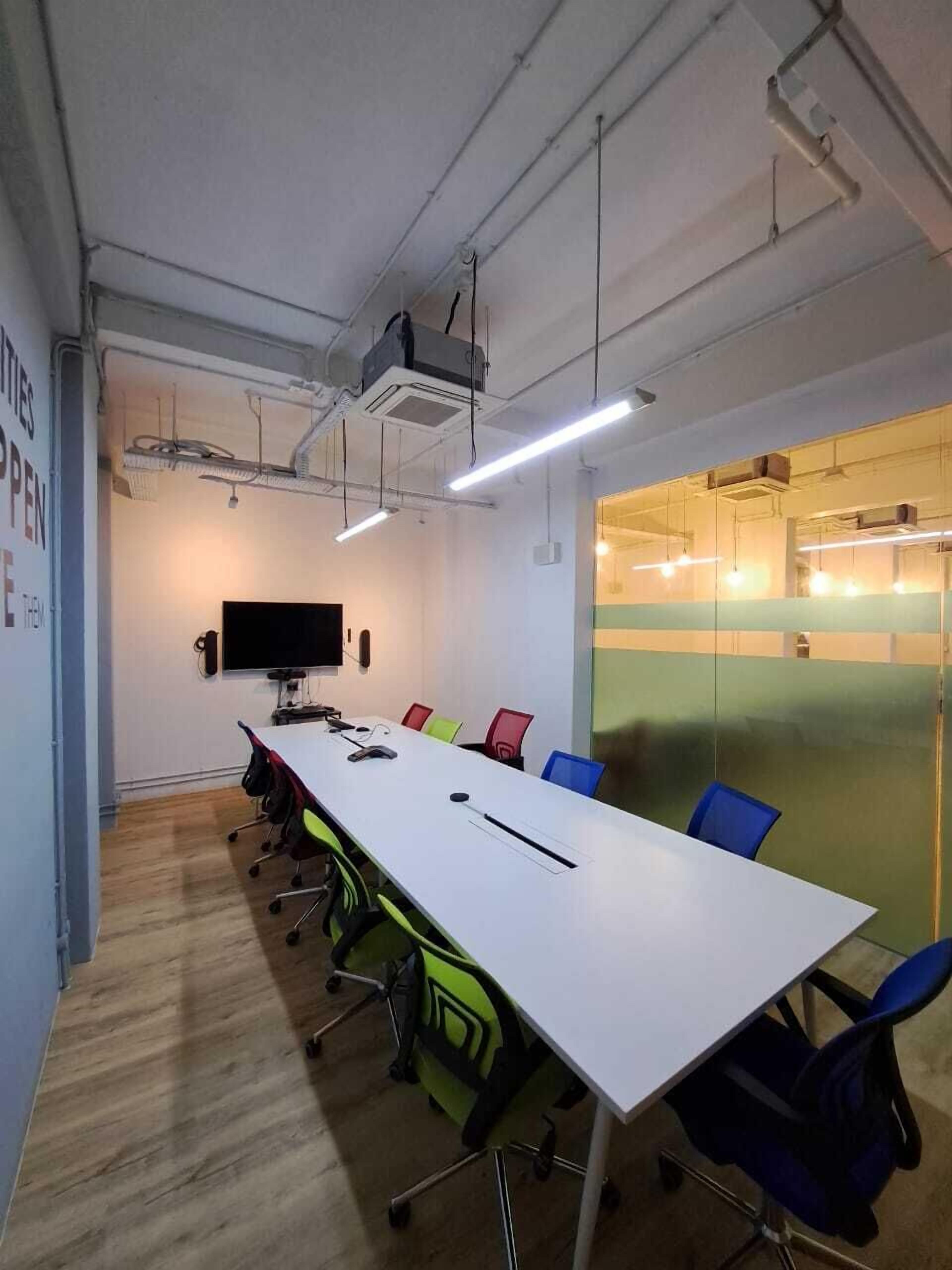 A conference room with a long white table, colorful chairs, a flat-screen TV, and a glass partition.