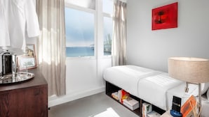 A modern hotel room with a white bed, a bedside table with a lamp, a view of the sea, and a red artwork on the wall. - Cooliv Waterfront (Singapore)