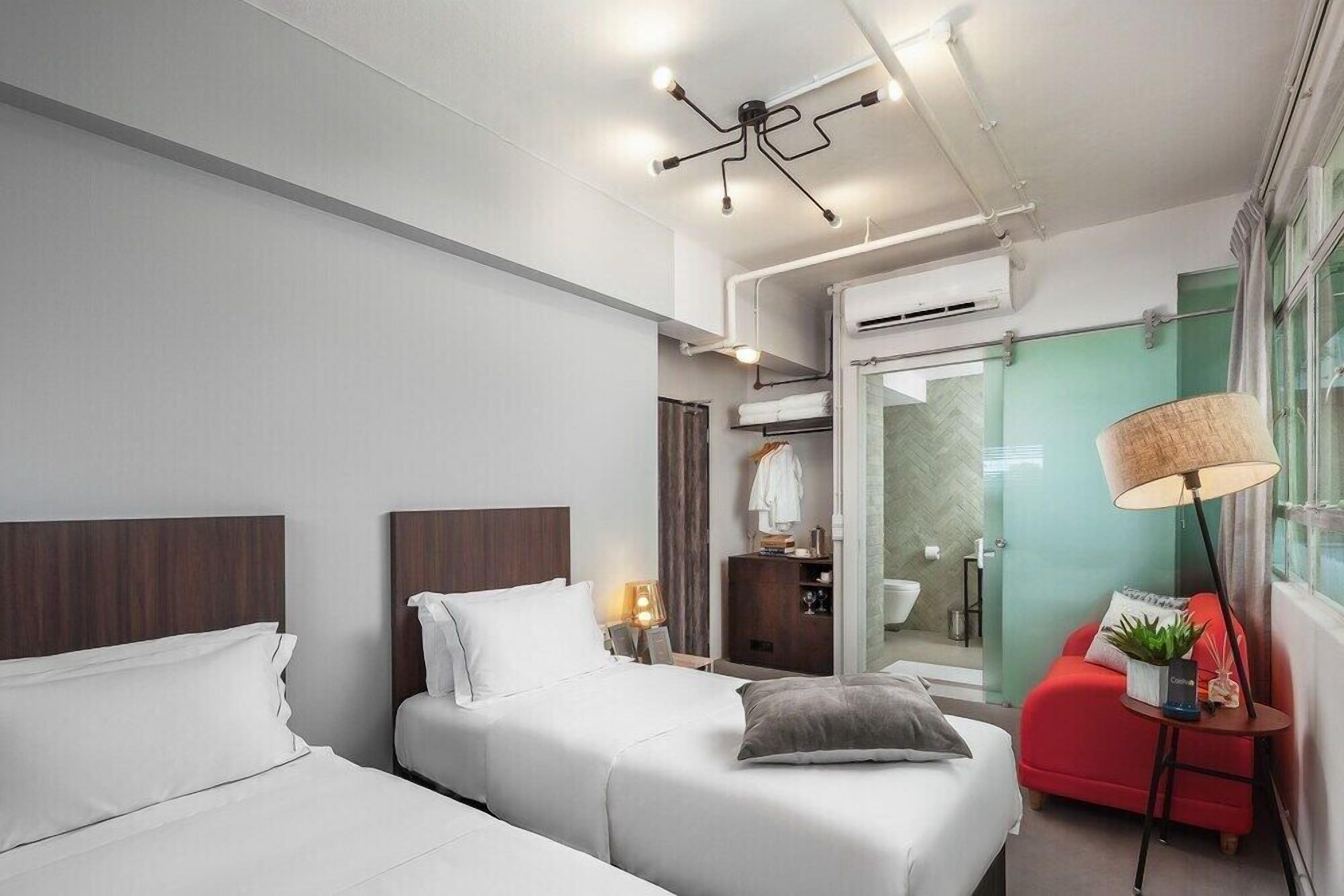 A modern hotel room with two beds, a red armchair, a lamp, and a glass shower enclosure.