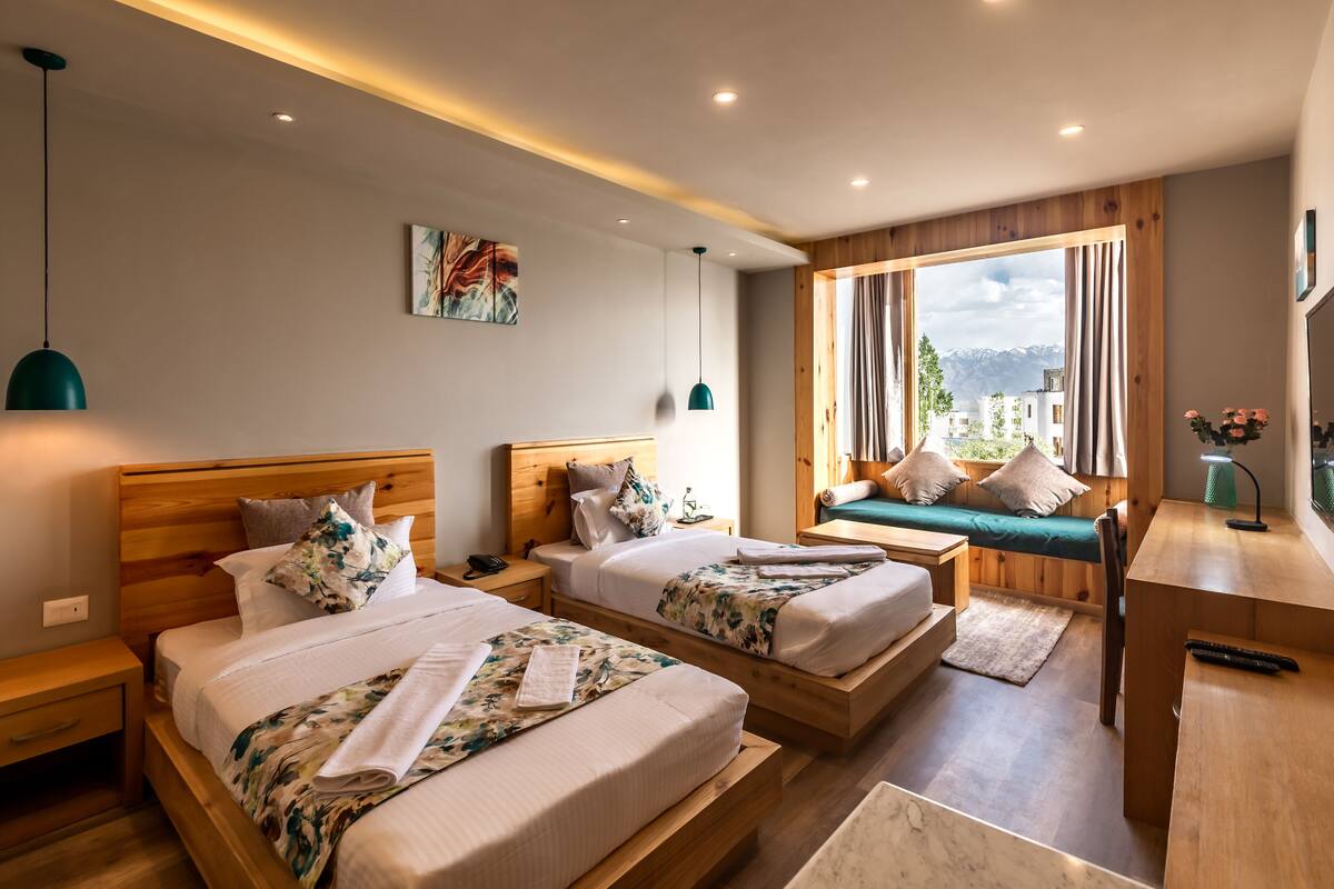 Quarto de hotel moderno com duas camas de madeira, uma escrivaninha e vista para montanhas através de amplas janelas.