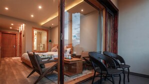 A hotel room with a wooden floor, a bed with a white headboard, a wooden desk with a chair, and a large window with a view of the sky. - The Bodhi Tree Ladakh (Leh)