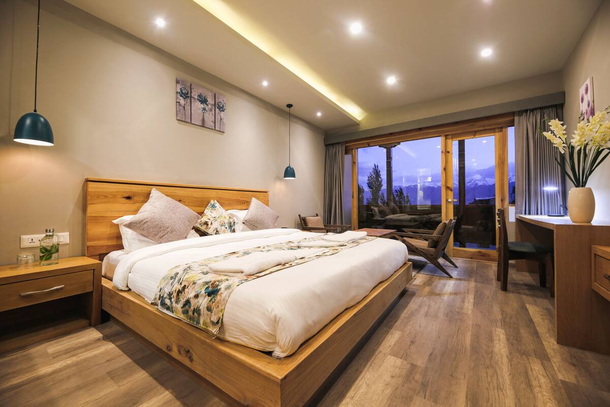 Quarto moderno com cama de madeira, mesas de cabeceira, escrivaninha e uma grande janela com vista para árvores e montanhas.