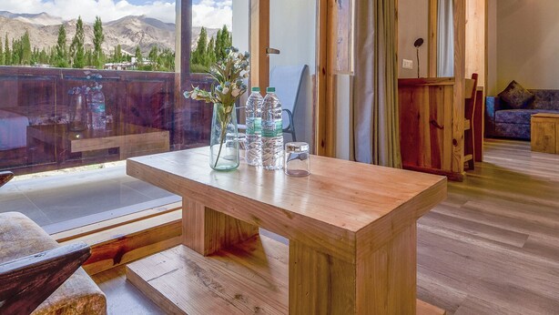 A wooden dining table with a vase and water bottles on it, a bench, and a view of mountains through a large window. - The Bodhi Tree Ladakh (Leh)