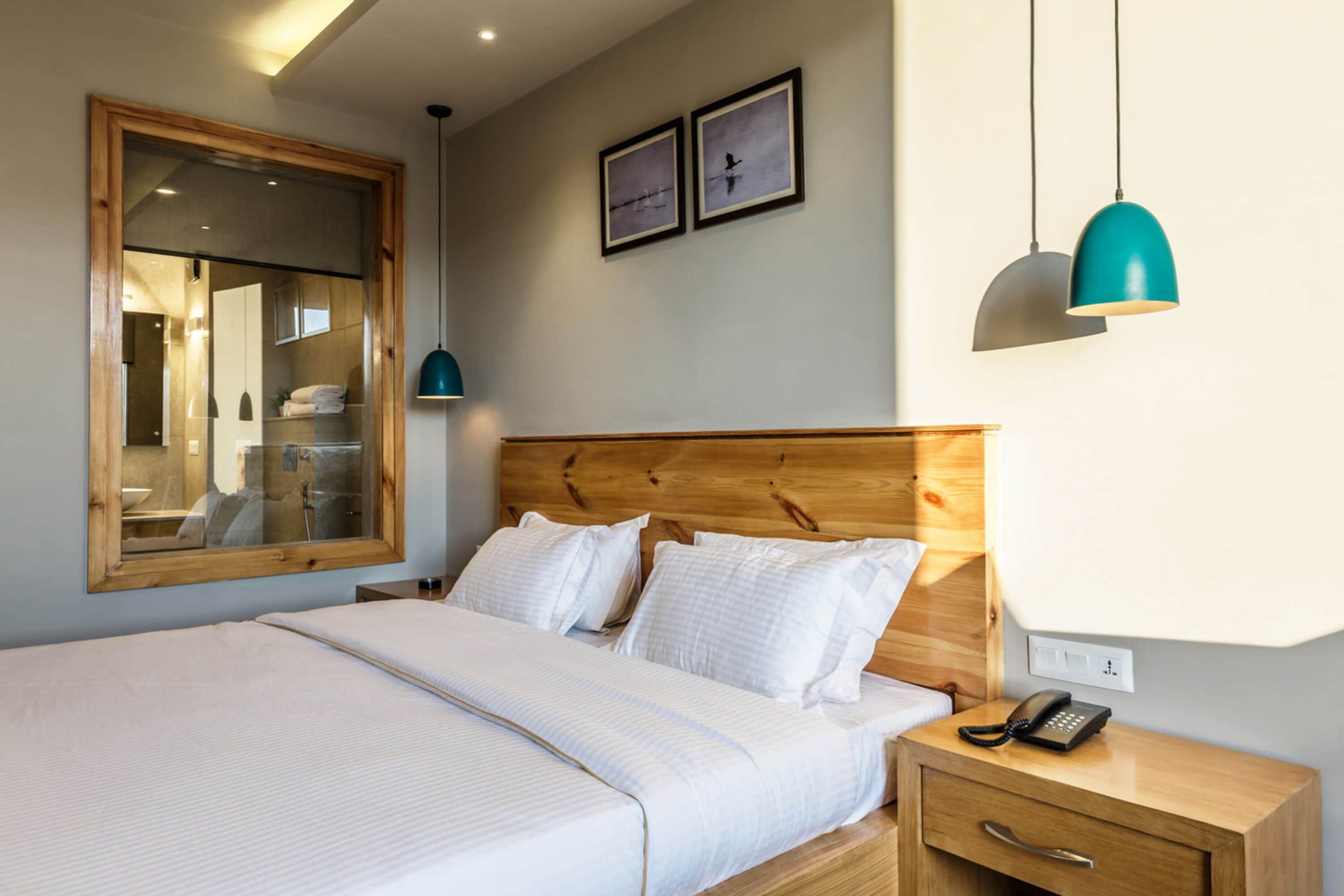A neatly made bed with white linens, a wooden headboard, a bedside table with a phone, and a mirror above the bed.