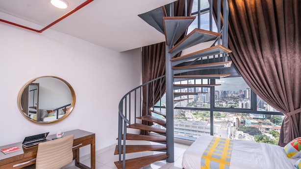Une chambre moderne avec un escalier en colimaçon, un bureau avec une chaise, un lit recouvert d’une couverture colorée et une grande fenêtre donnant sur la ville.