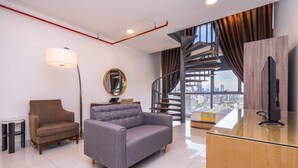 Un salon moderne avec un canapé gris, un fauteuil marron, un miroir rond, une lampe sur pied, un téléviseur posé sur un meuble en bois et une vue sur la ville à travers de grandes fenêtres.