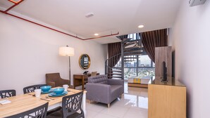 Un salon moderne avec un canapé, une table à manger et des chaises. Un escalier mène à l’étage supérieur, et de grandes fenêtres offrent une vue sur la ville.