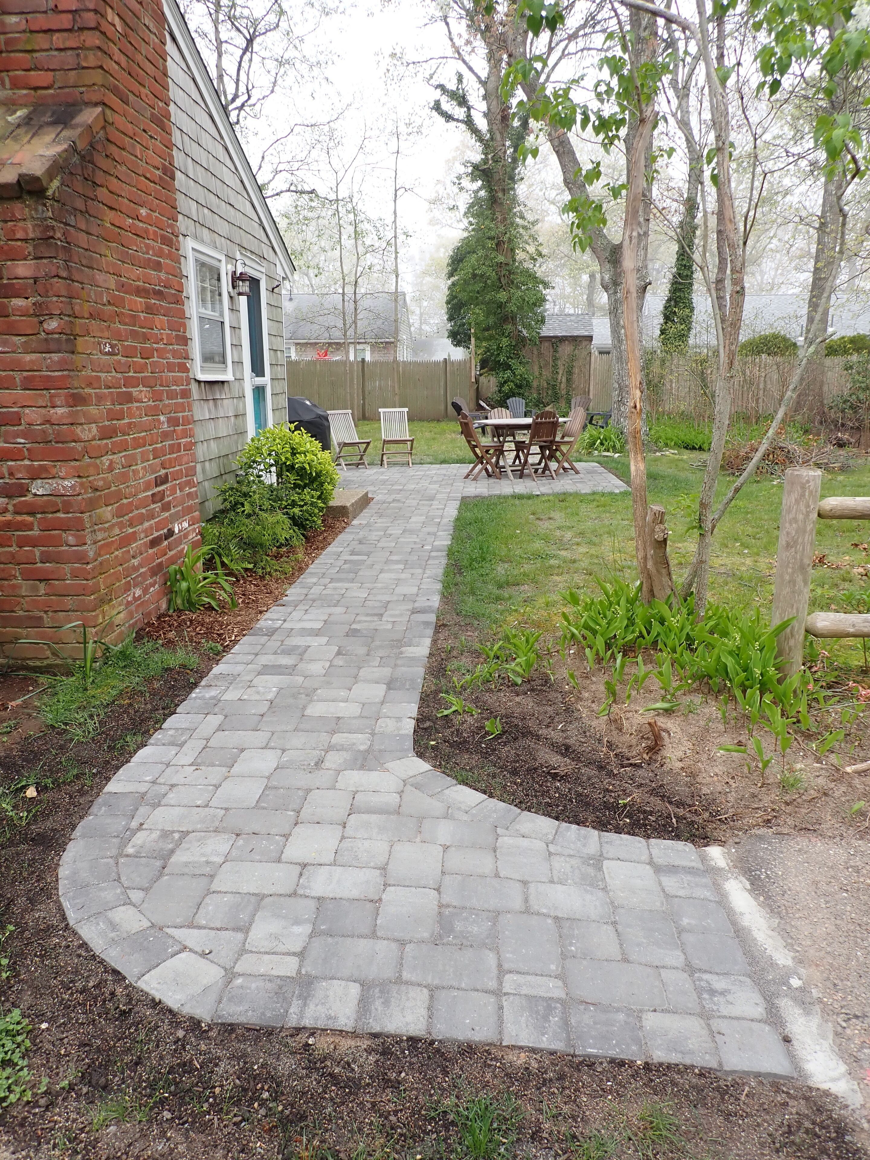 A brick pathway leading to a patio area with a table and chairs, surrounded by greenery and a wooden fence.