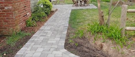 Un sendero de ladrillos que conduce a un patio con una mesa y sillas, rodeado de follaje y una valla de madera.