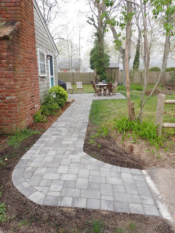 A brick pathway leading to a patio area with a table and chairs, surrounded by greenery and a wooden fence.