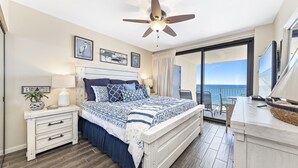 A bedroom with a bed, a nightstand, a lamp, a ceiling fan, and a view of the ocean.