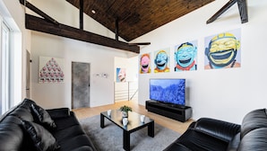 Una sala de estar moderna con sofás de cuero negro, una mesa de centro y un televisor sobre un mueble. Hay cuadros coloridos en la pared y una escalera al fondo.