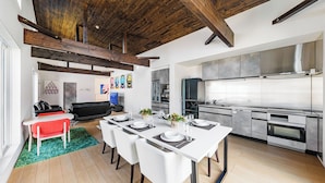 Una cocina moderna con electrodomésticos de acero inoxidable, un comedor con sillas blancas y una mesa para cuatro, y una sala de estar con sofá y televisor.