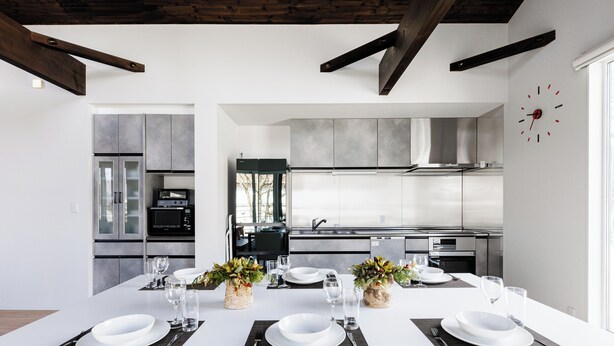 Una cocina moderna con electrodomésticos de acero inoxidable, una zona de comedor con mesa y sillas blancas, y un reloj en la pared.