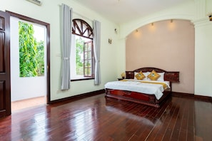 Un dormitorio con piso de madera, una cama grande con cabecera, una ventana con cortinas y vistas al exterior.