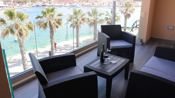 Un balcón con mobiliario de mimbre, una mesa con una botella y vasos, y vistas a un puerto deportivo con barcos y palmeras.