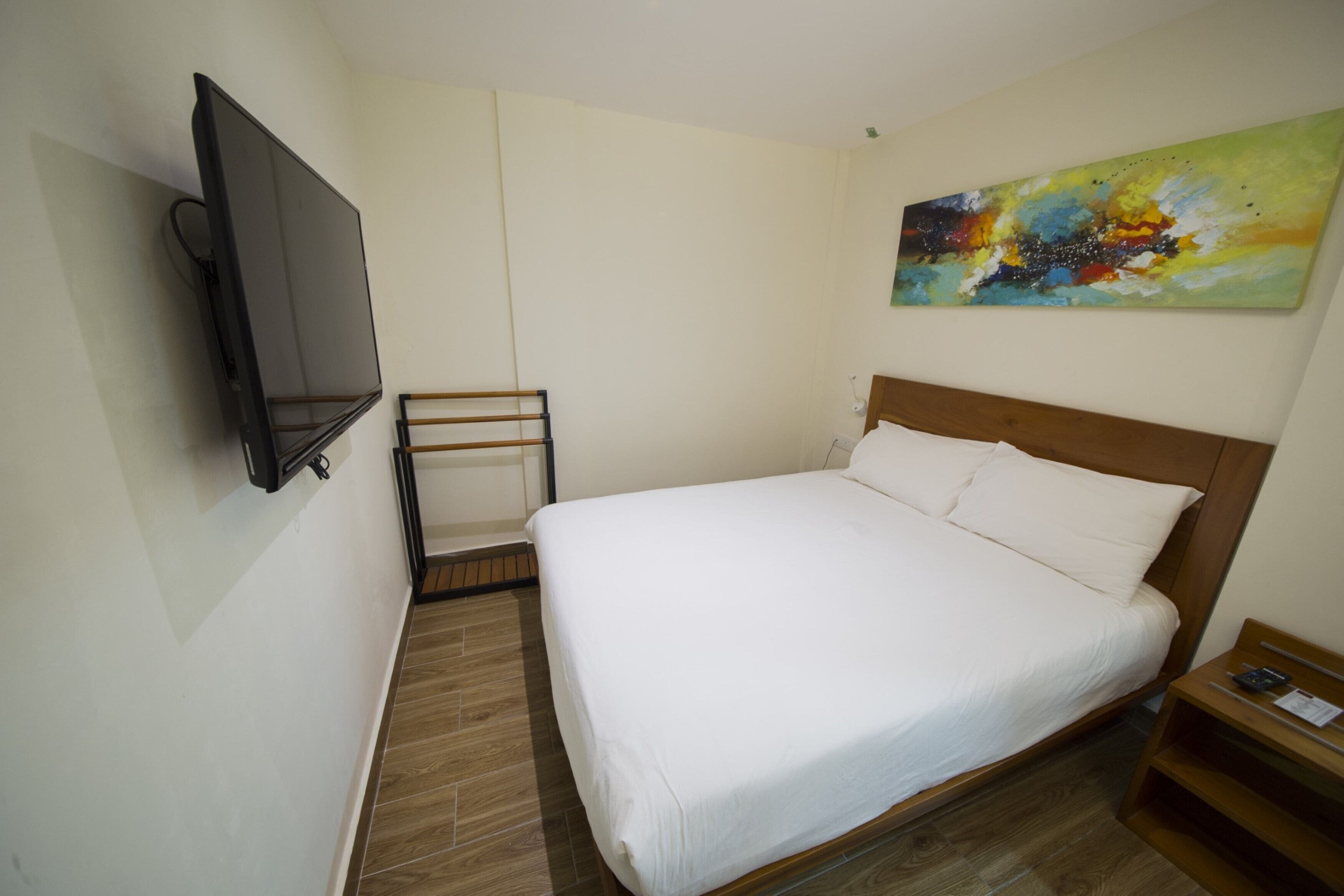 A hotel room with a bed, a TV mounted on the wall, a painting on the wall, and a wooden nightstand.