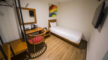 Una habitación de hotel compacta con una cama individual, un escritorio con una silla, una mesita pequeña, un espejo y un cuadro colorido en la pared.