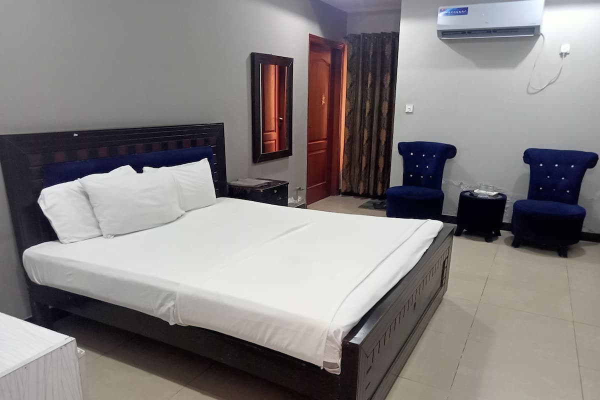 A hotel room with a large bed, two armchairs, a small table, a wardrobe, and an air conditioning unit.