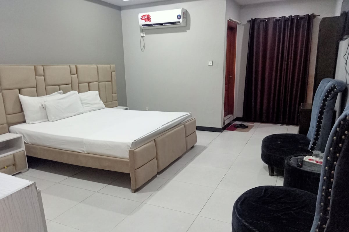 A hotel room with a large bed, a bedside table, a chair, and an air conditioning unit on the wall.