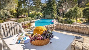 Un tavolo con un cesto di uva e un libro, una sedia bianca e una piscina sullo sfondo.