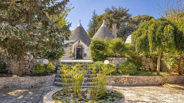Un edificio in pietra con tetti a cono, immerso in un giardino con una fontana circolare e diverse piante.