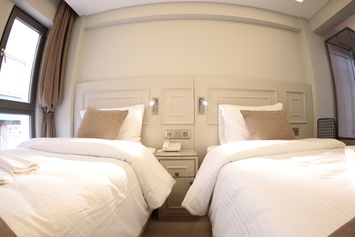 Quarto de hotel com duas camas, mesa de cabeceira com telefone e janela com cortinas.