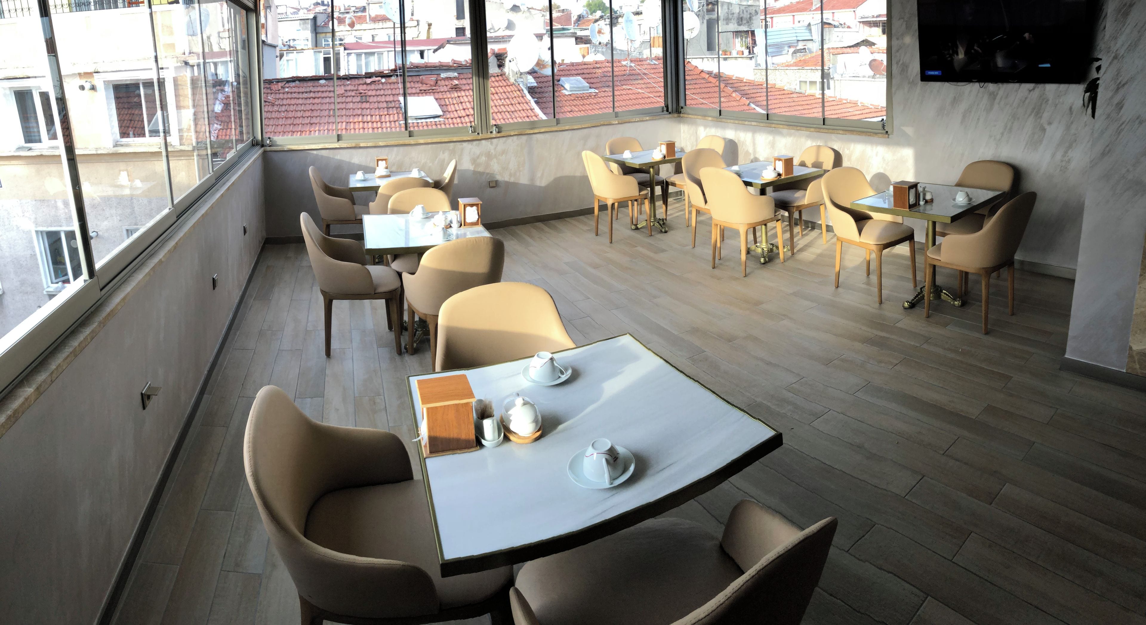 A modern dining area with multiple tables and chairs, large windows offering a view of the cityscape, and a flat-screen TV mounted on the wall.
