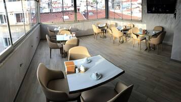 A modern dining area with multiple tables and chairs, large windows offering a view of the cityscape, and a flat-screen TV mounted on the wall.