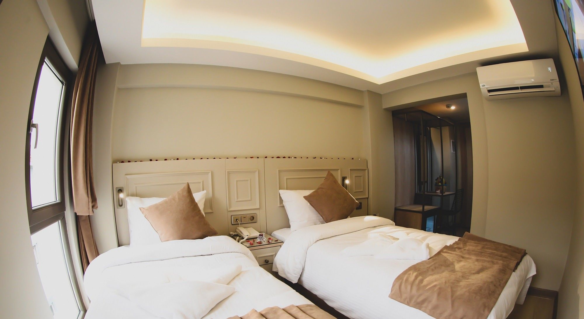 A hotel room with two beds, a bedside table with a lamp, an air conditioning unit, and a window with curtains.