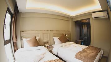 A hotel room with two beds, a bedside table with a lamp, an air conditioning unit, and a window with curtains.