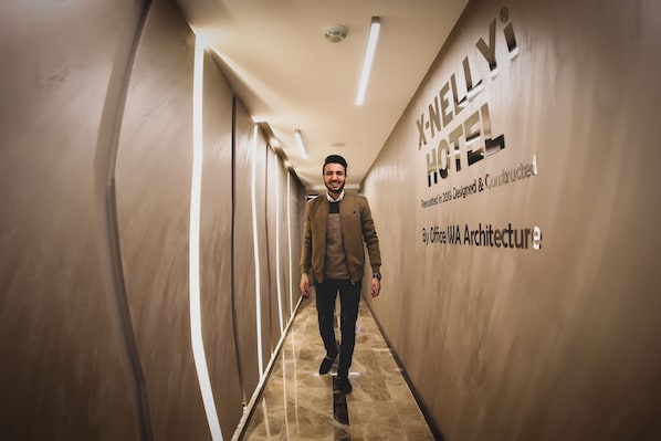 Um homem caminhando por um corredor com uma placa que diz "X-NELLY HOTEL" e menciona "Reformado em 2018, projetado e construído pela Office WA Architecture".
