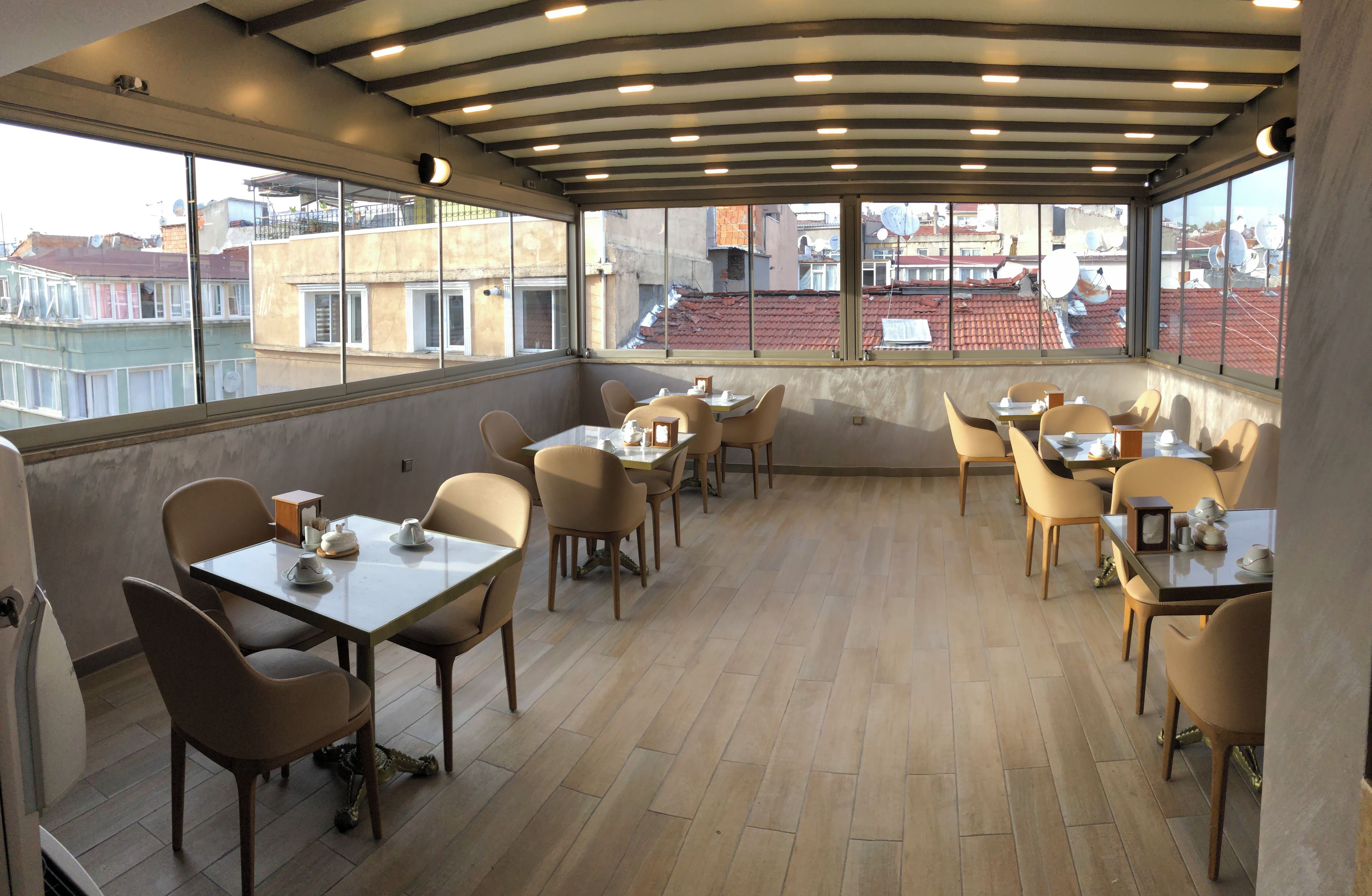 A rooftop restaurant with tables and chairs, large windows, and a view of buildings.