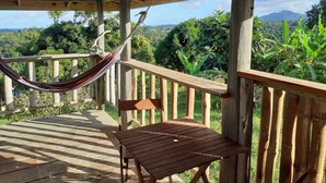 Una terraza de madera con una hamaca y una mesa, con vistas a un paisaje exuberante y montañas a lo lejos.