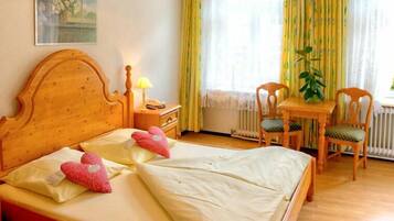 A hotel room with two beds, a wooden headboard, a bedside table with a lamp, a chair, and a window with curtains.