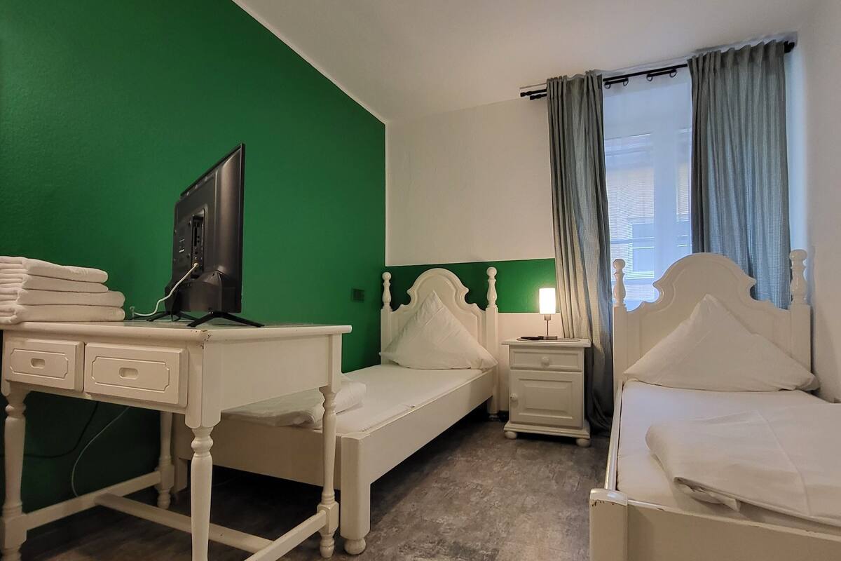 Une chambre d’hôtel avec un mur vert, deux lits blancs, un bureau avec une télévision et une table de chevet avec une lampe.