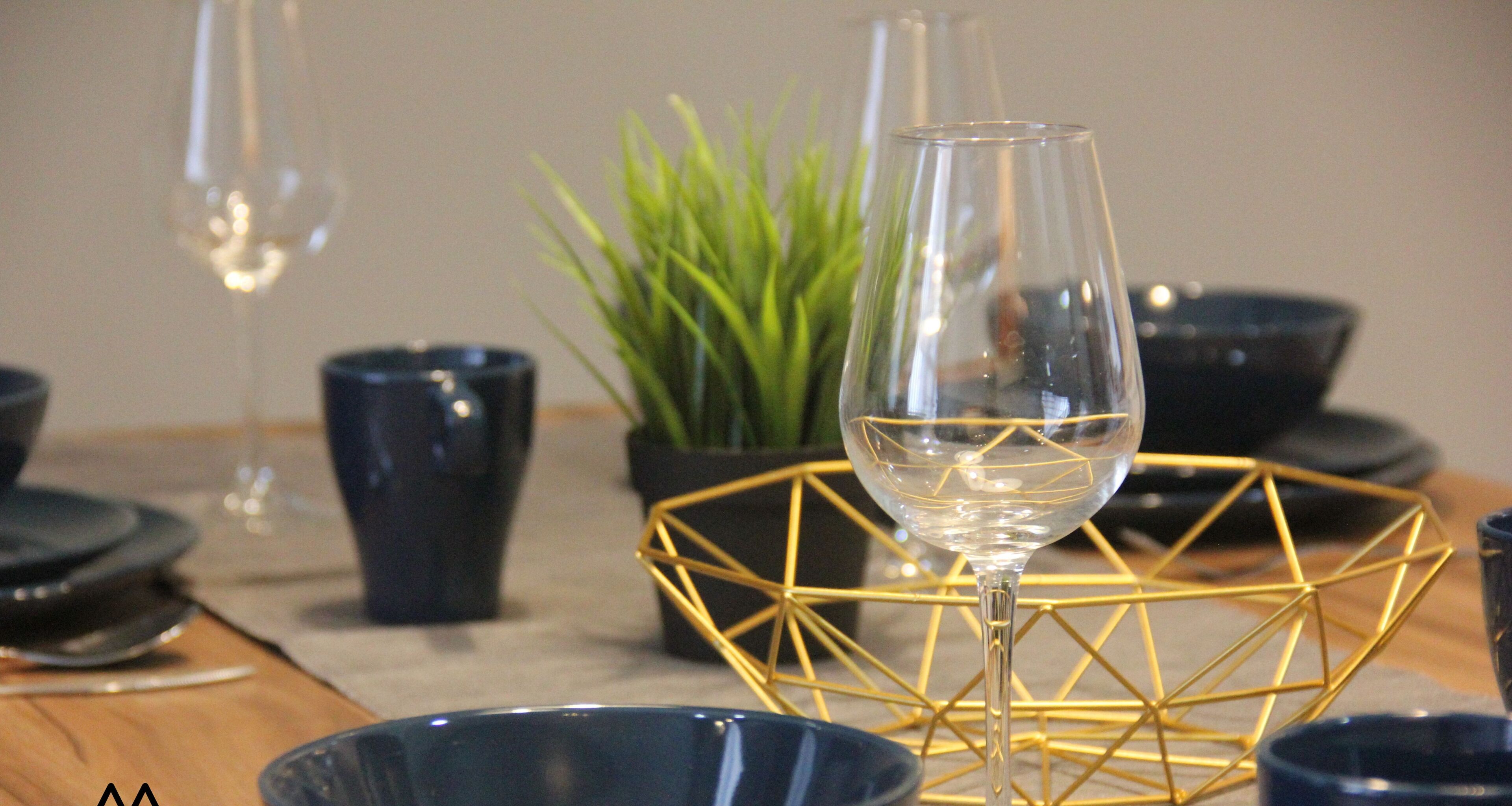 Una mesa de comedor con una copa de vino, una planta en maceta y un centro de mesa geométrico dorado.