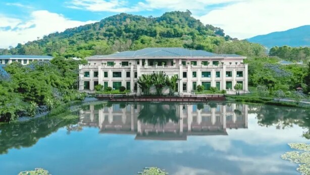 Un grande edificio con specchio d'acqua di fronte, circondato da vegetazione e colline.