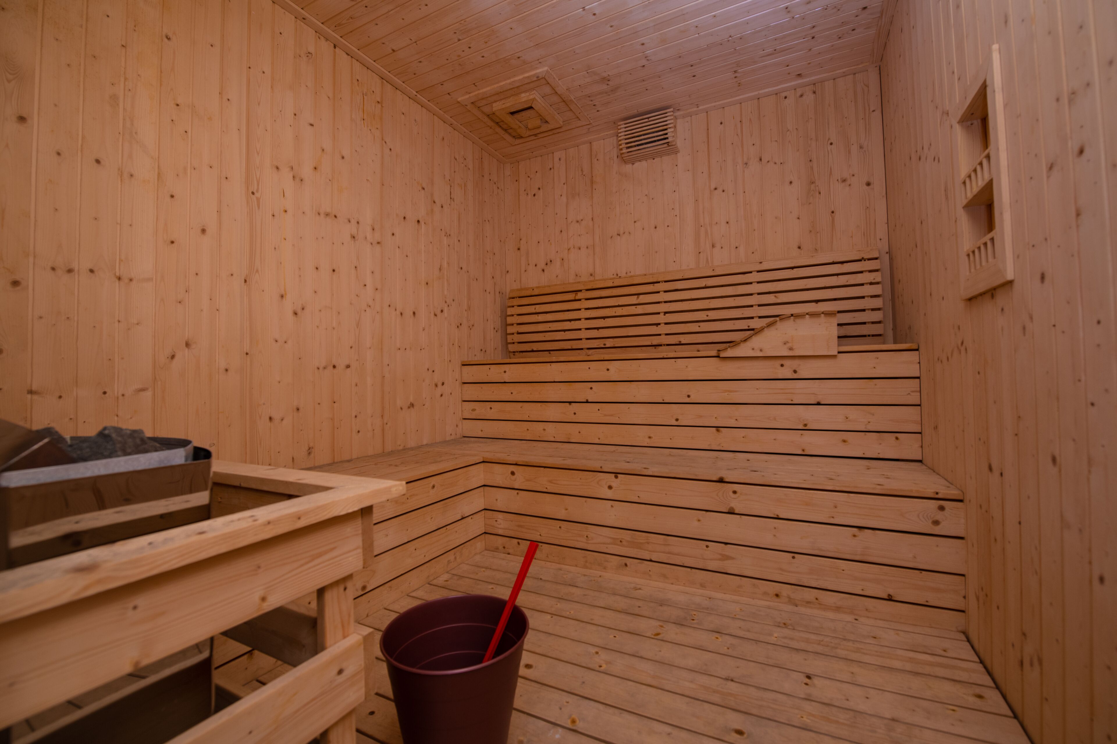 A sauna with wooden walls and benches, a bucket with a red handle, and a ventilation vent on the ceiling.