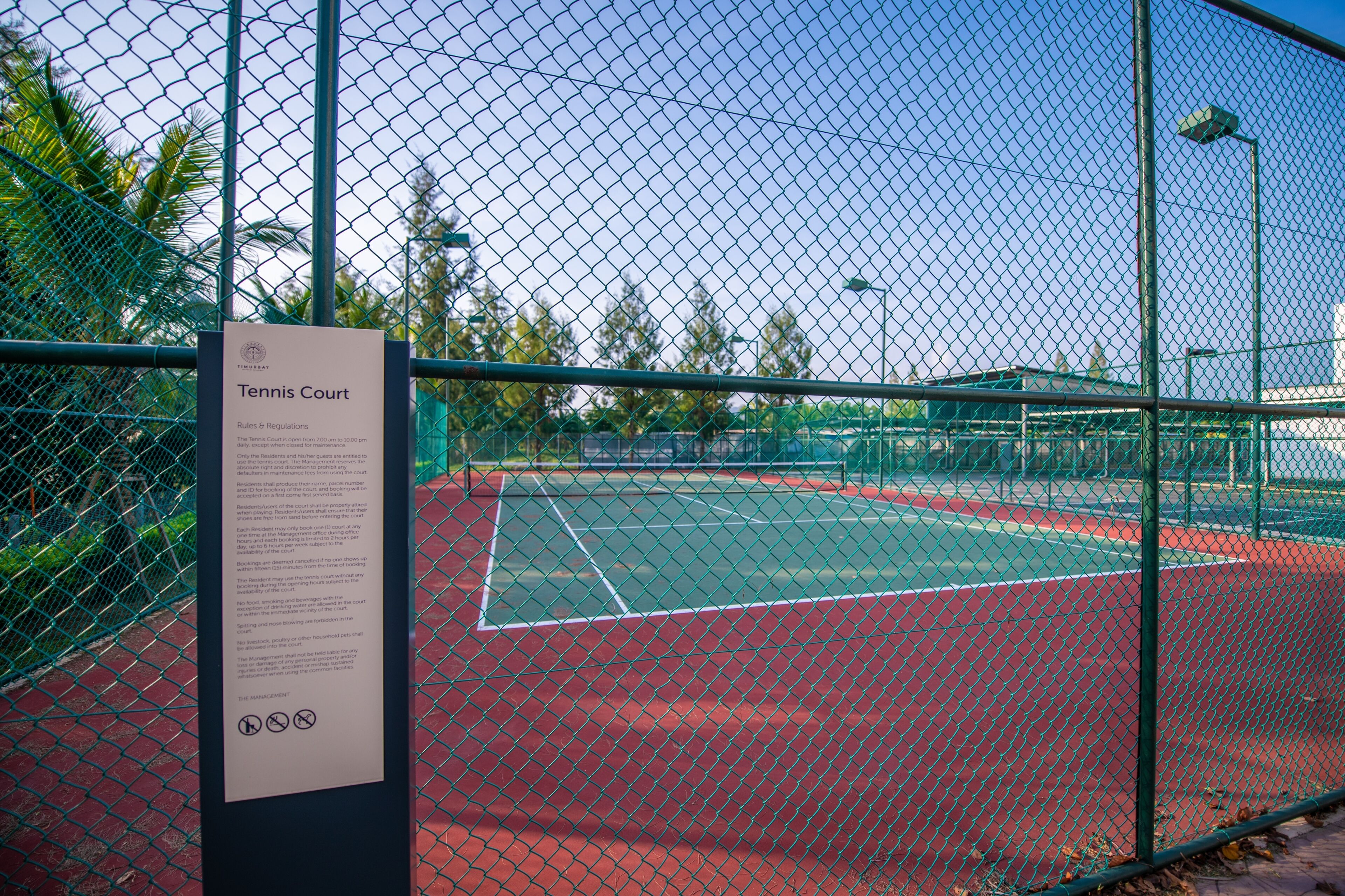 Cancha de tenis rodeada por una cerca de malla metálica, con un cartel que detalla normas y reglamentos.