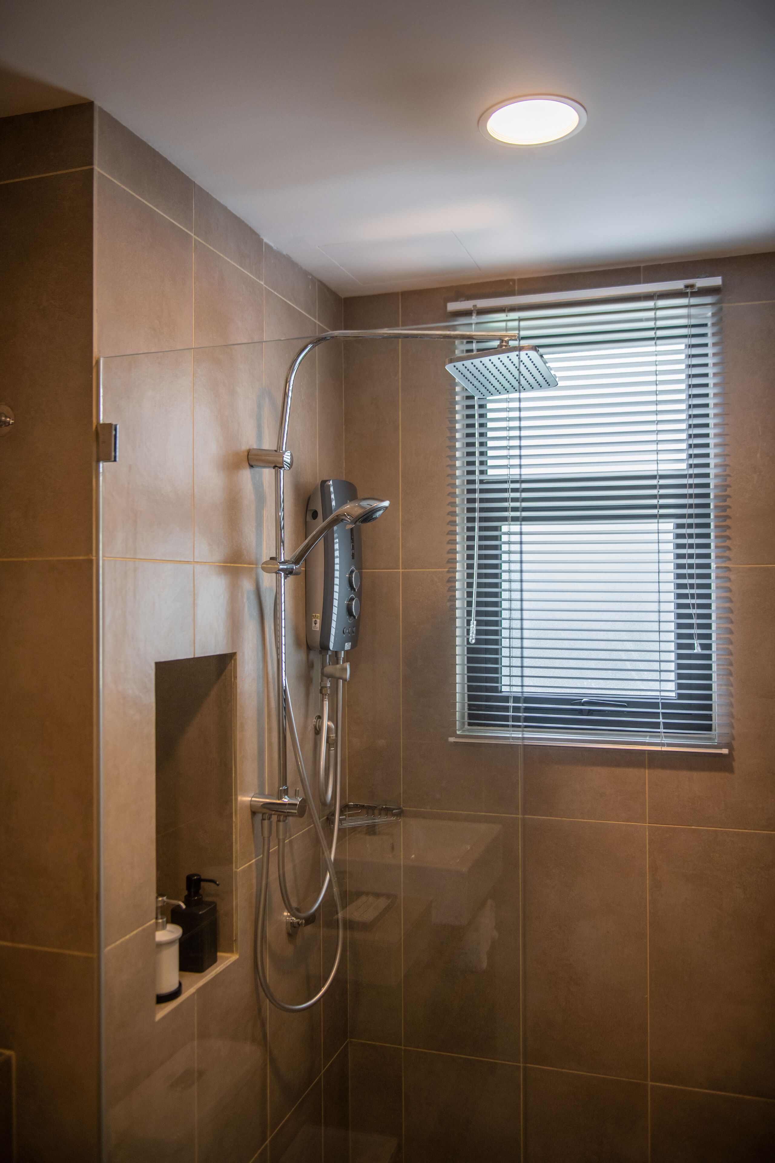 A modern shower with a handheld showerhead and a wall-mounted faucet, a window with blinds, and a small shelf with toiletries.